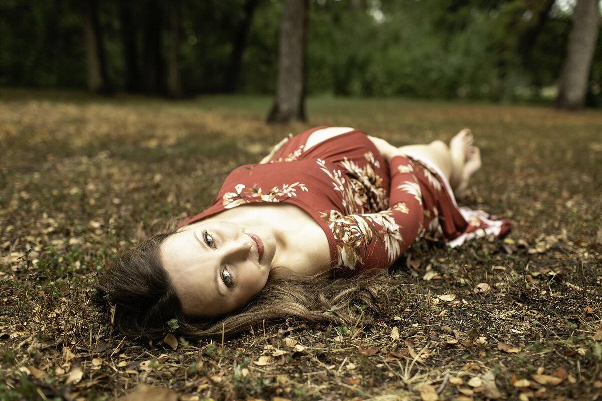A serene maternity photo of the expectant mom in a red dress, lying on a soft grassy patch at Trinity Park, gently cradling her belly as the sun filters through the trees, captured by Chunky Monkey Photography.