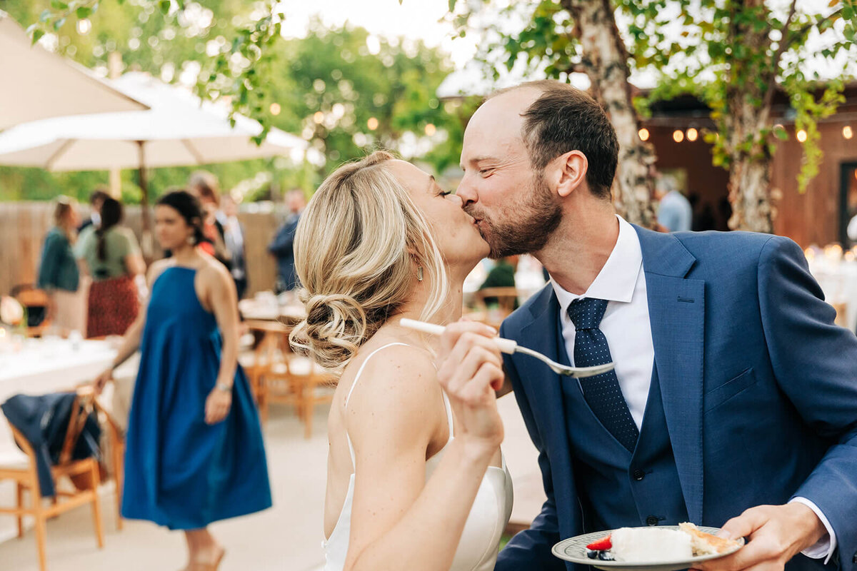 A bride and groom kissing while holding a slice of wedding cake.