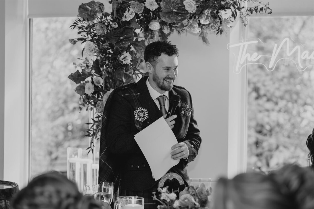 A groom stands in front of a large floral display and holds a piece of paper. He smiles and deliver a speech.