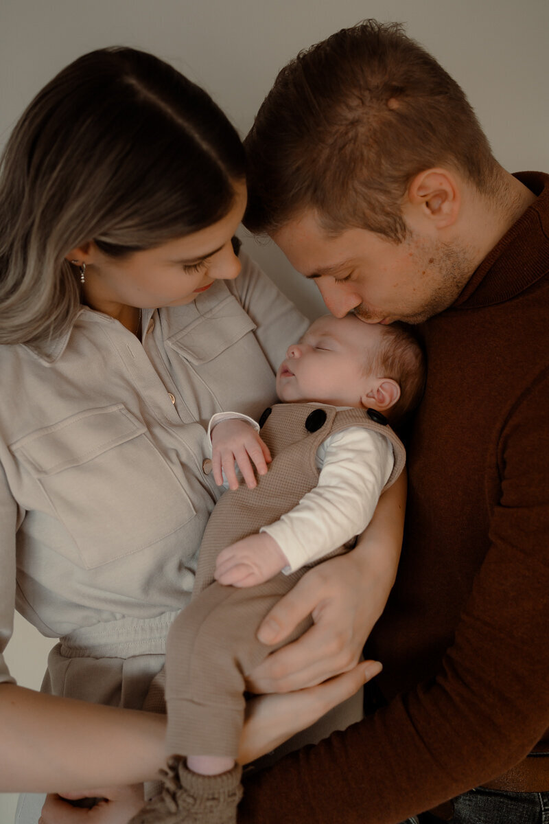 moeder heeft zoontje op haar arm terwijl vader een kusje op het hoofd van de baby geeft