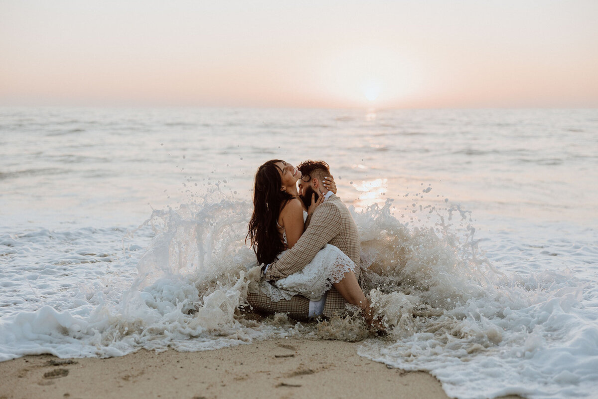 couple sitting on the beach with an ocean wave crashing into them