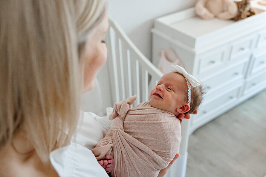 Baby smiles while being held by her mom with a crib in background.