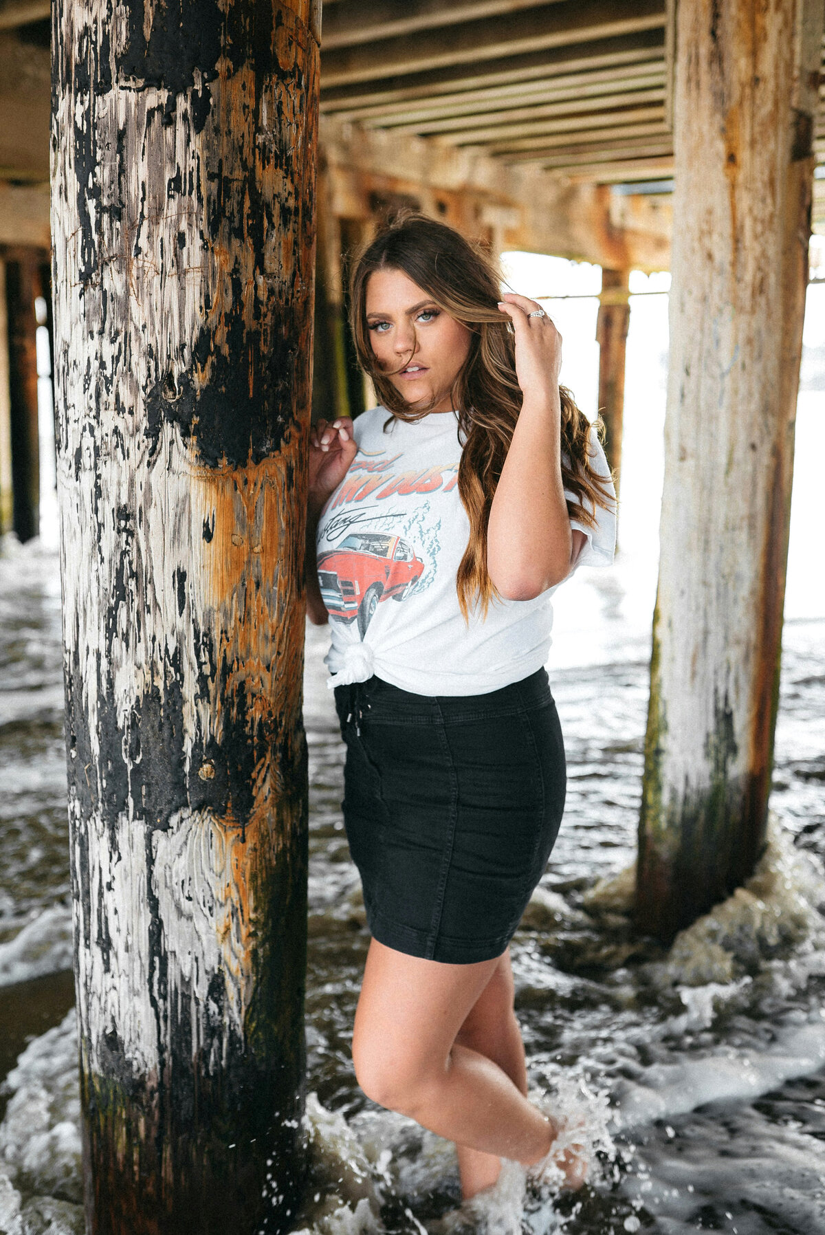 High school senior standing under a pier, wearing a t-shirt and jean skirt looking confidently at the camera.