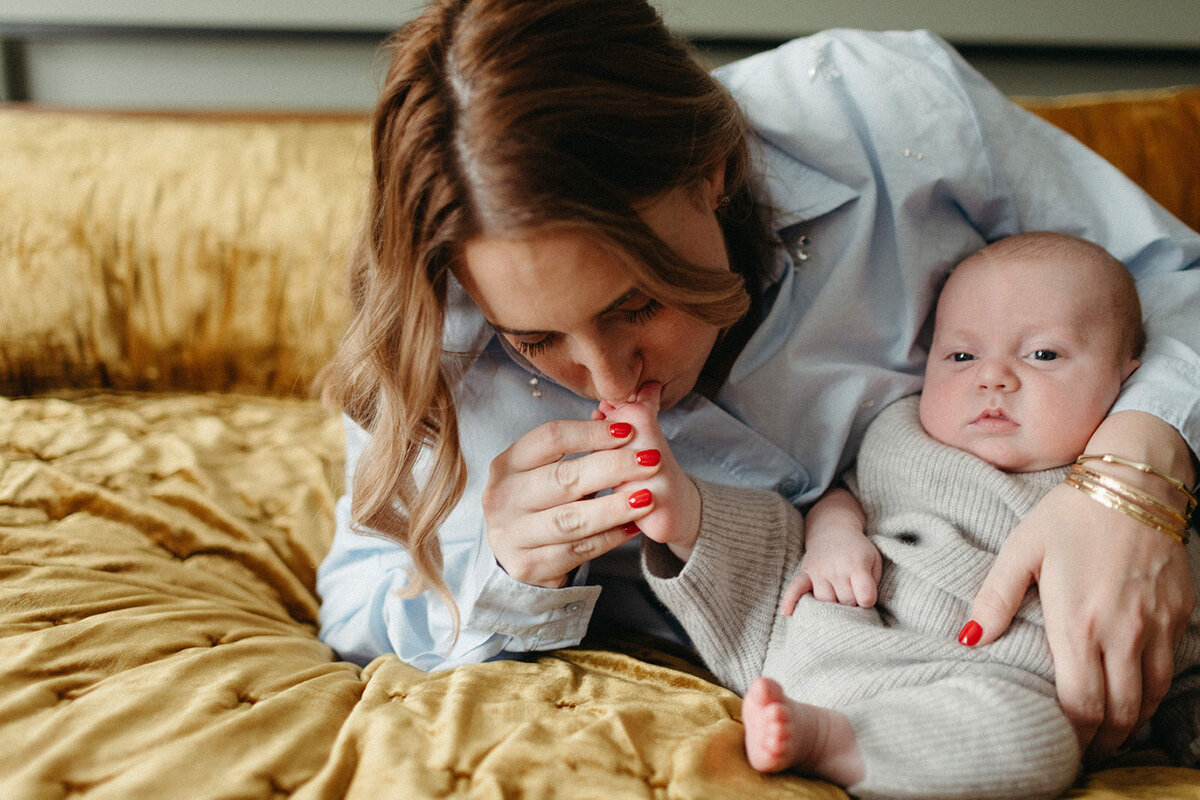In a heartwarming moment captured by a talented Oshawa newborn photographer, a woman with red nails tenderly kisses a baby's hand, cradling the child on a mustard-colored quilt. The baby looks adorable in their gray knit outfit.