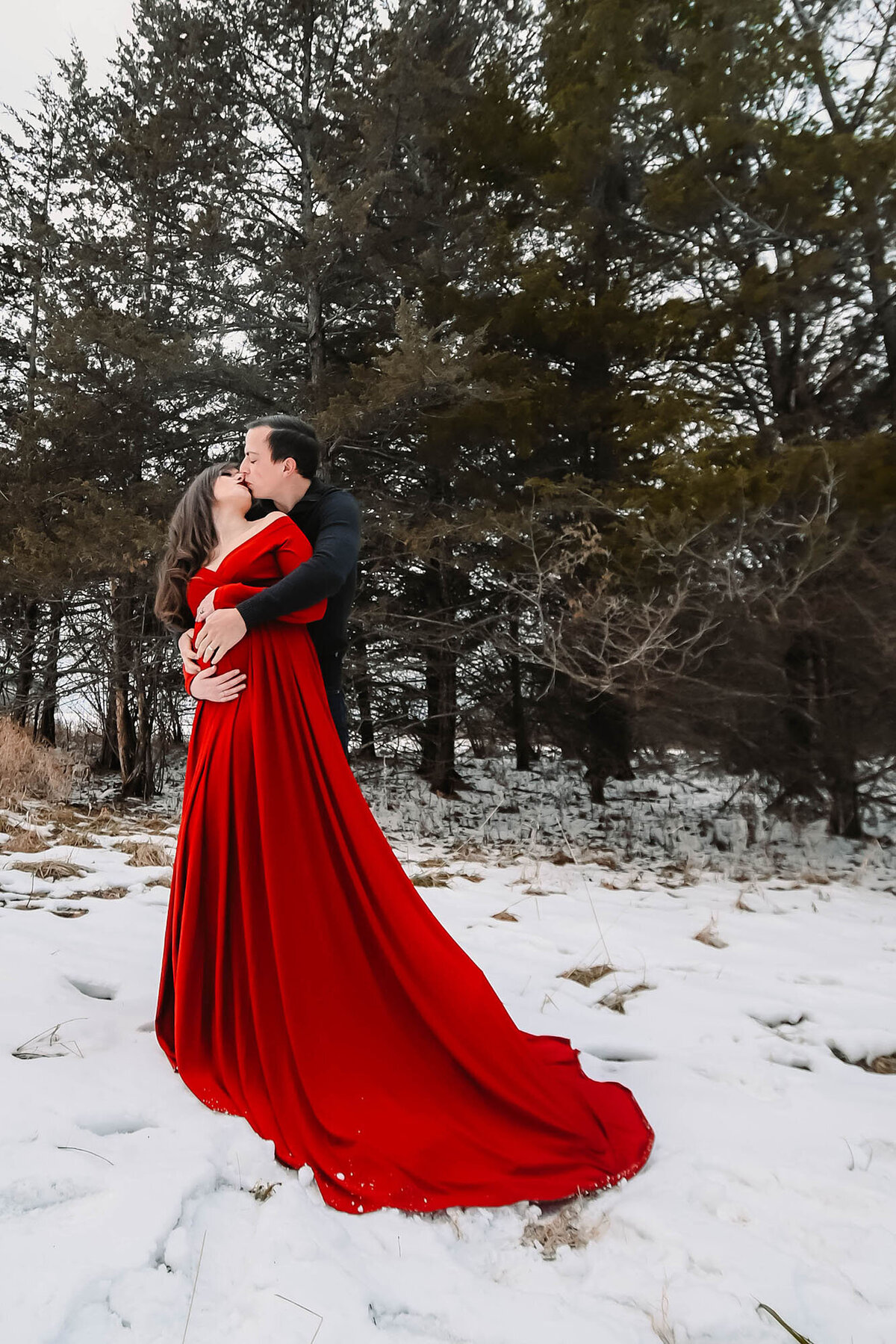 A happy pregnant couple kisses in the snow in a black shirt and red maternity gown