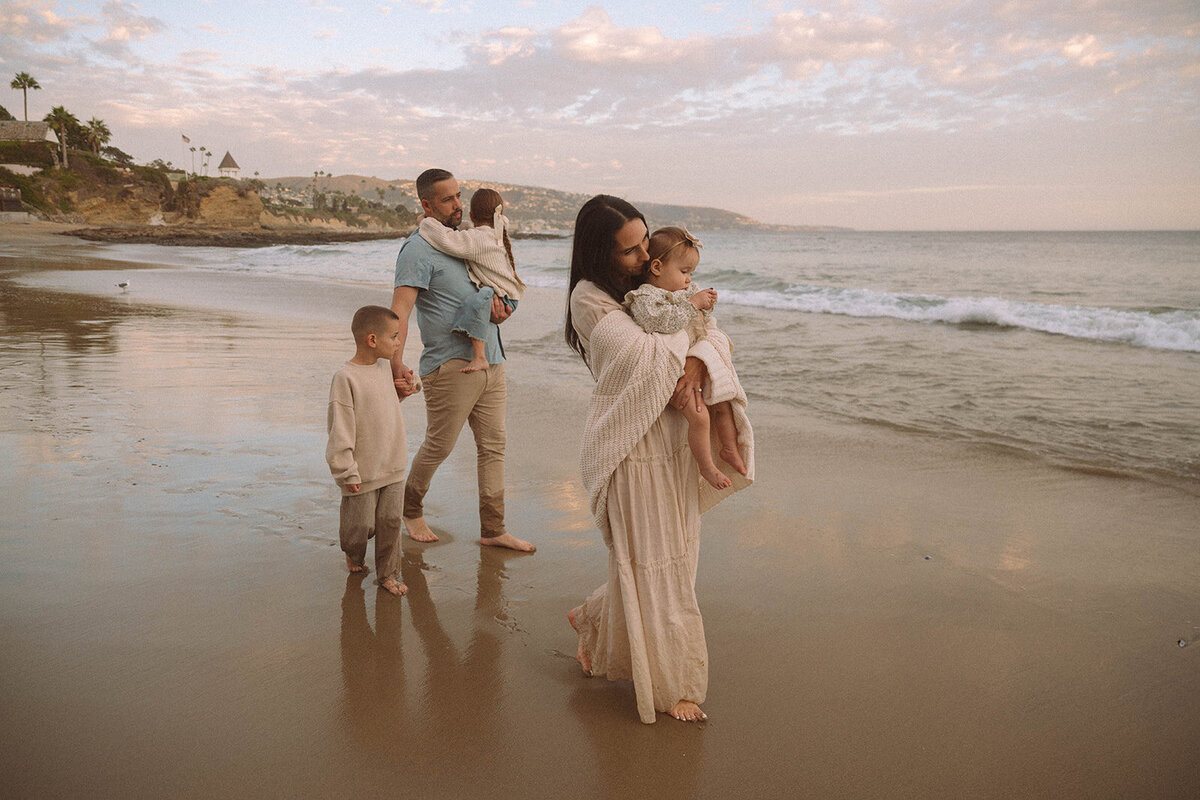 Family walking along the shore at Shaw’s Cove in Laguna Beach during sunset, with parents carrying their kids and soft pastel skies overhead — photographed by Orange County family photographer Maria Alcantara.
