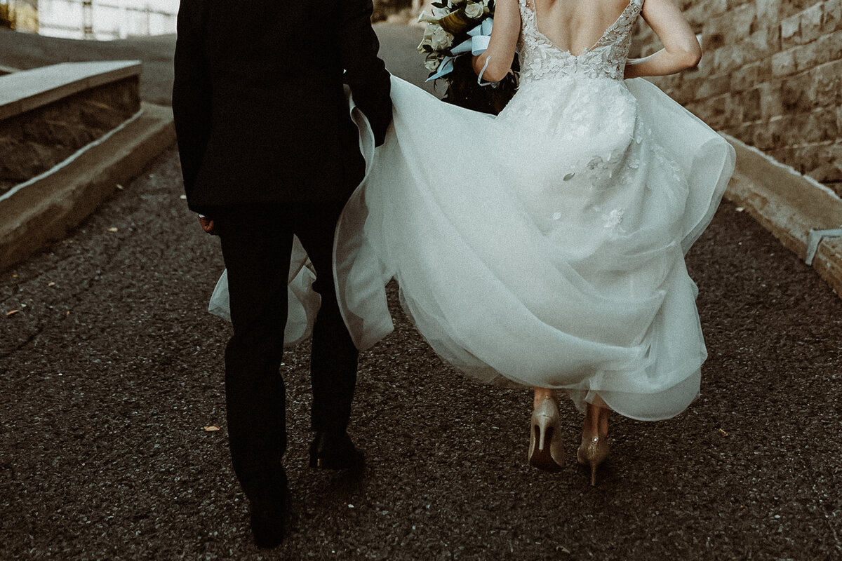 groom holding his brides dress as they walk away