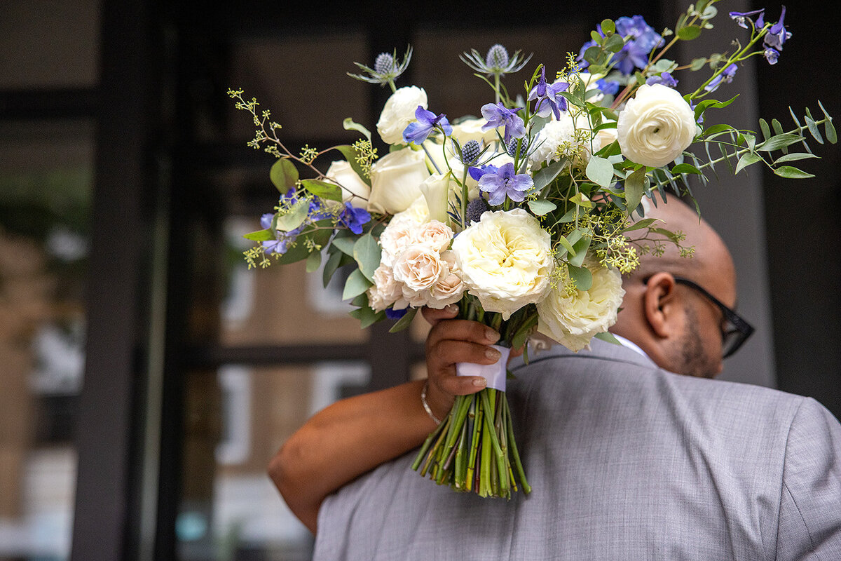 Bride holding a lush bouquet of white roses, spray roses, ranunculus, blue delphiniums, and greenery, ready for a wedding event.