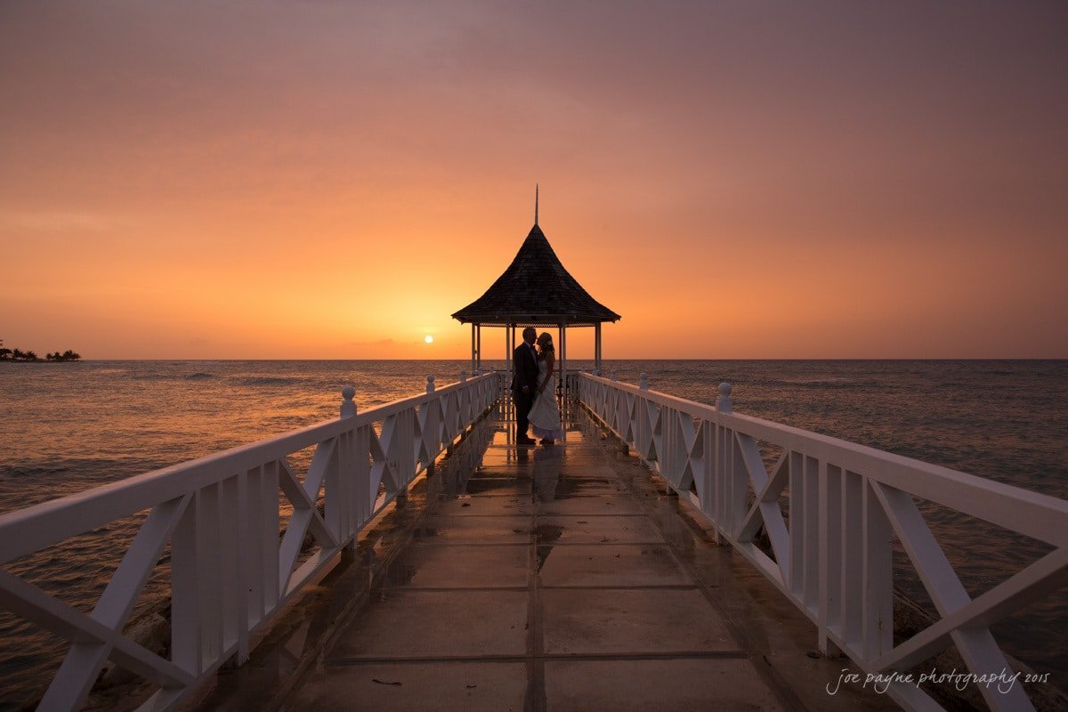 A wedding couple standing at the end of a pier at sunset.