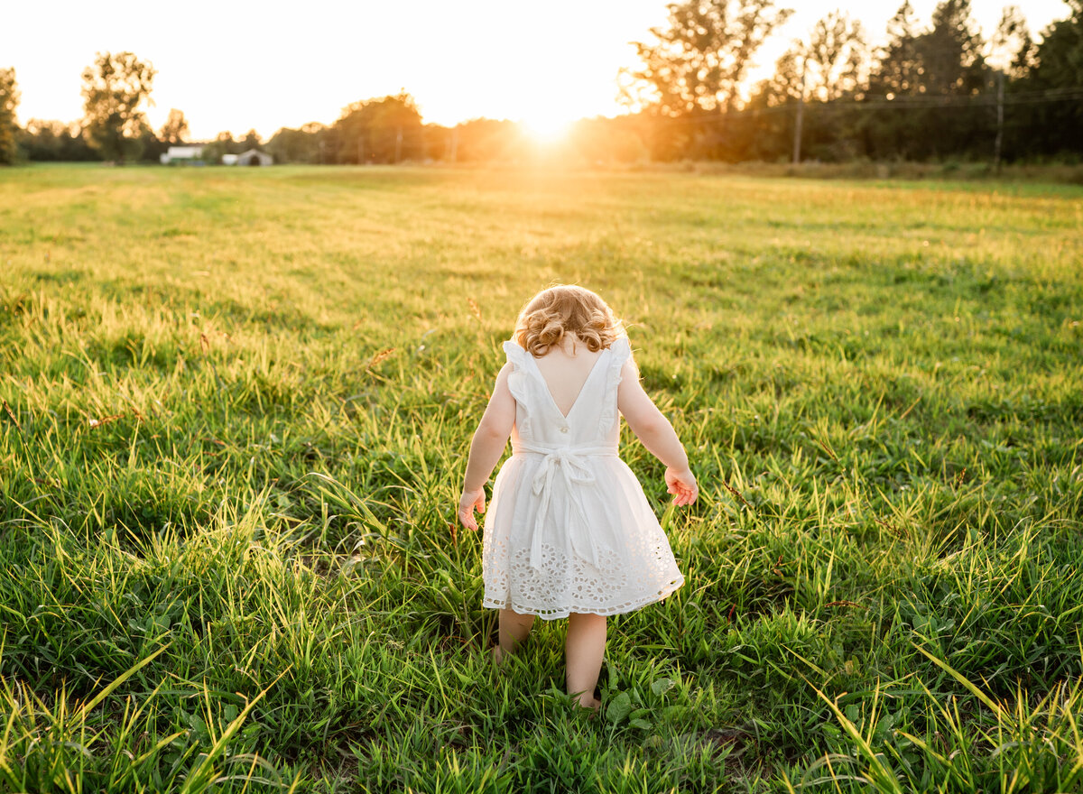 A young child in a white dress walking through a grassy field at sunset