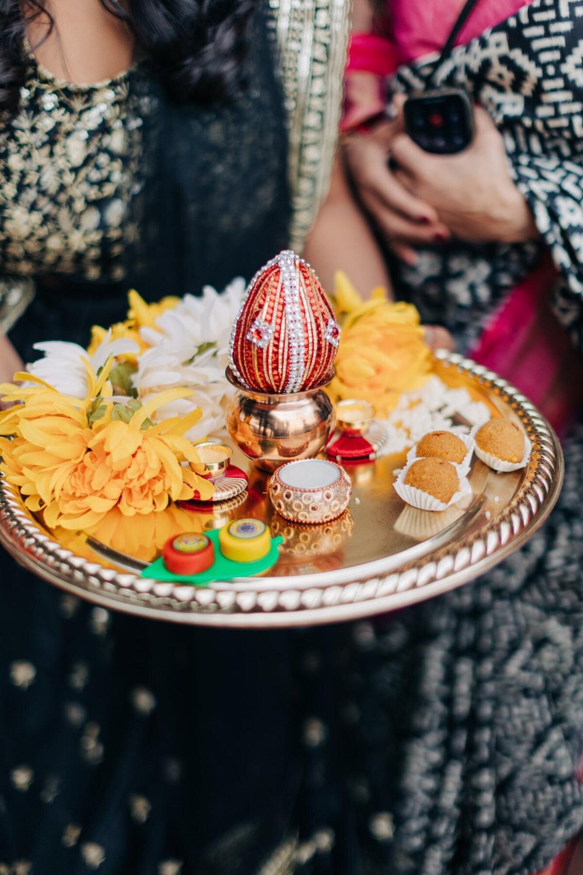 A decorative tray held by a woman, displaying flowers, a small ornate pot, and ritualistic items, possibly at an indian cultural or religious ceremony.