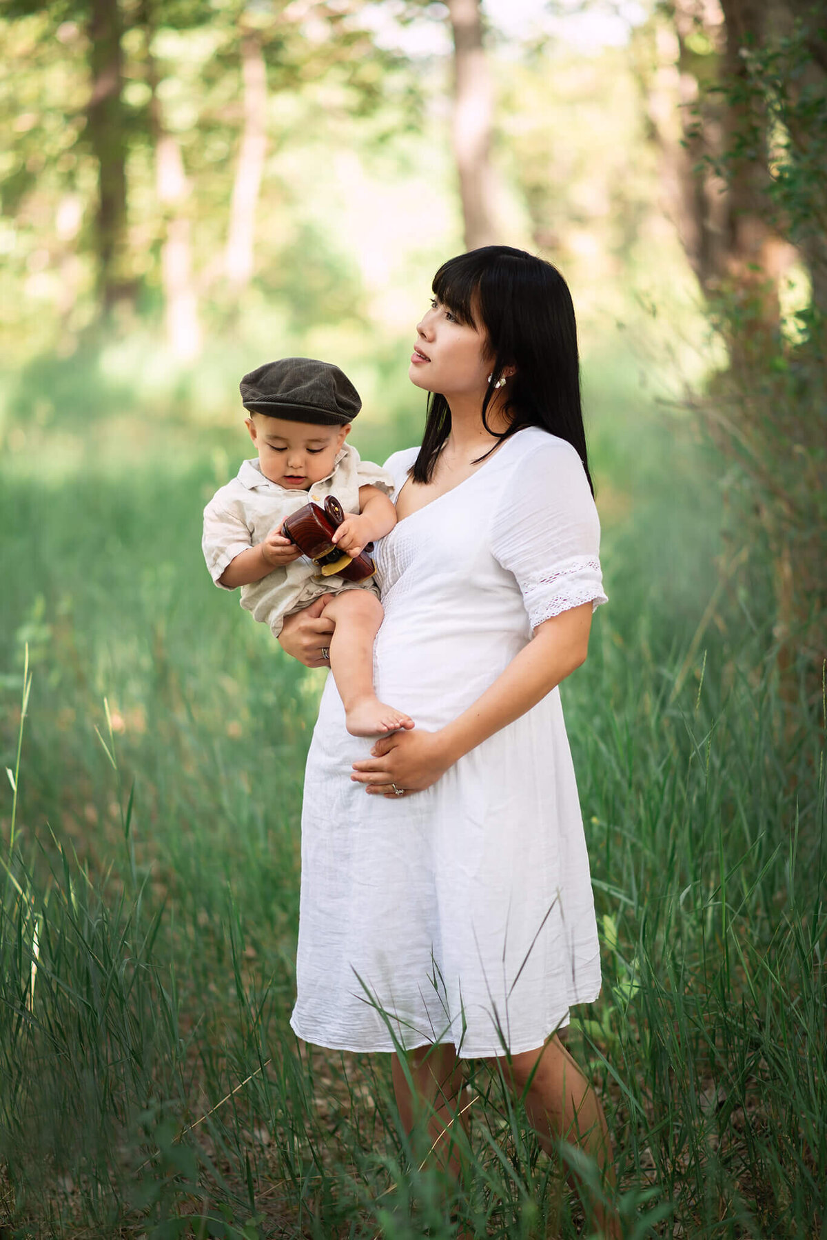 A woman wearing a white dress with long black hair cradles her pregnant belly while carrying a young toddler. She looks off into the distance while the young boy looks at the camera.  Brittany Danielle takes this photo.