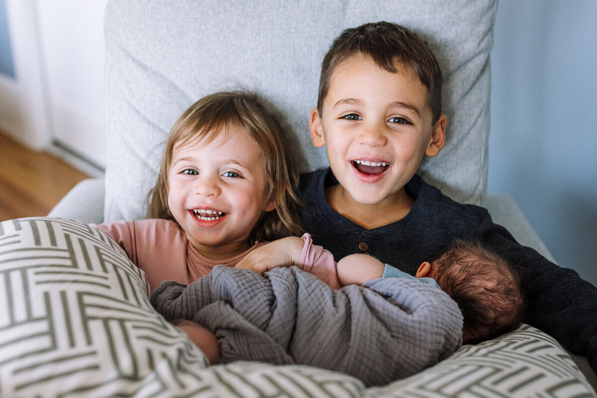 borther and sister sitting in chair looking at camera and smiling while holding swaddled newborn propped on patterned pillow