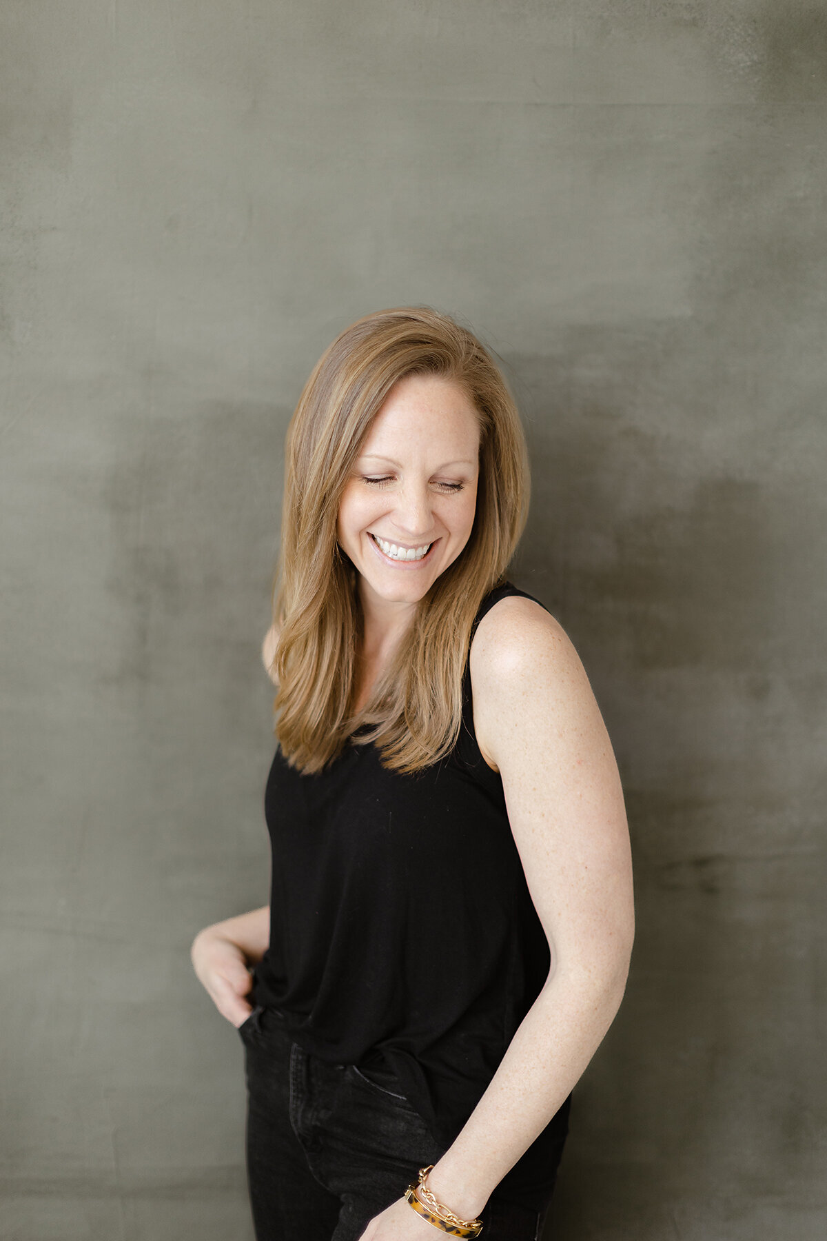 Professional headshot photo of a DFW business woman leaning against the wall in a Dallas photography studio as she has one hand in her pocket looking down at the floor.