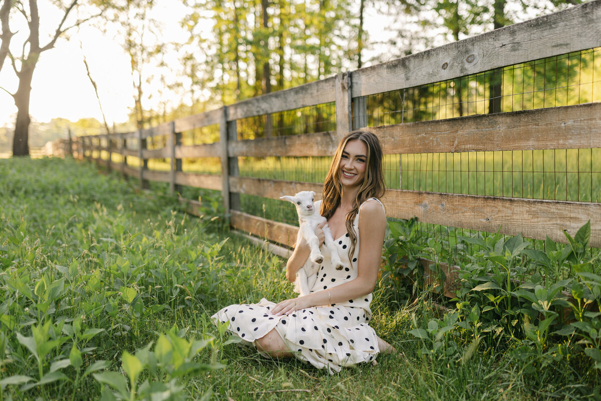 Young woman in a polka dot dress holding a baby goat during golden hour at a vintage family farm. Greenville SC branding photograph by Dani Lacey Photographs.