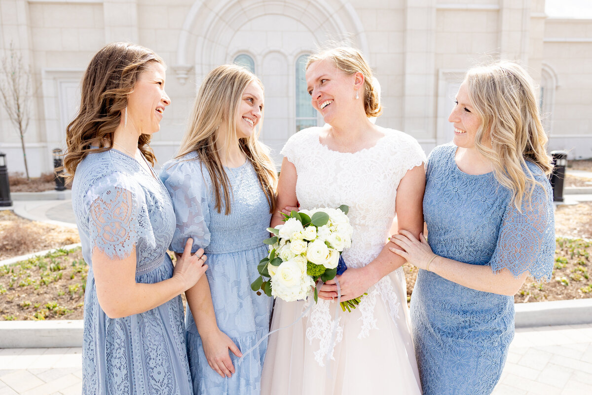 The bride stands with her three bridesmaids, all smiling warmly at each other in a candid, joyful moment.