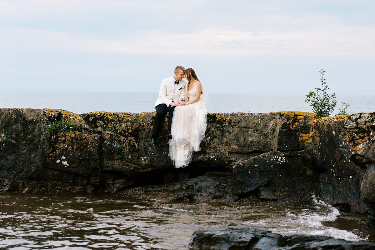Bride and groom sitting on the rocks of North Shore with their heads touching.