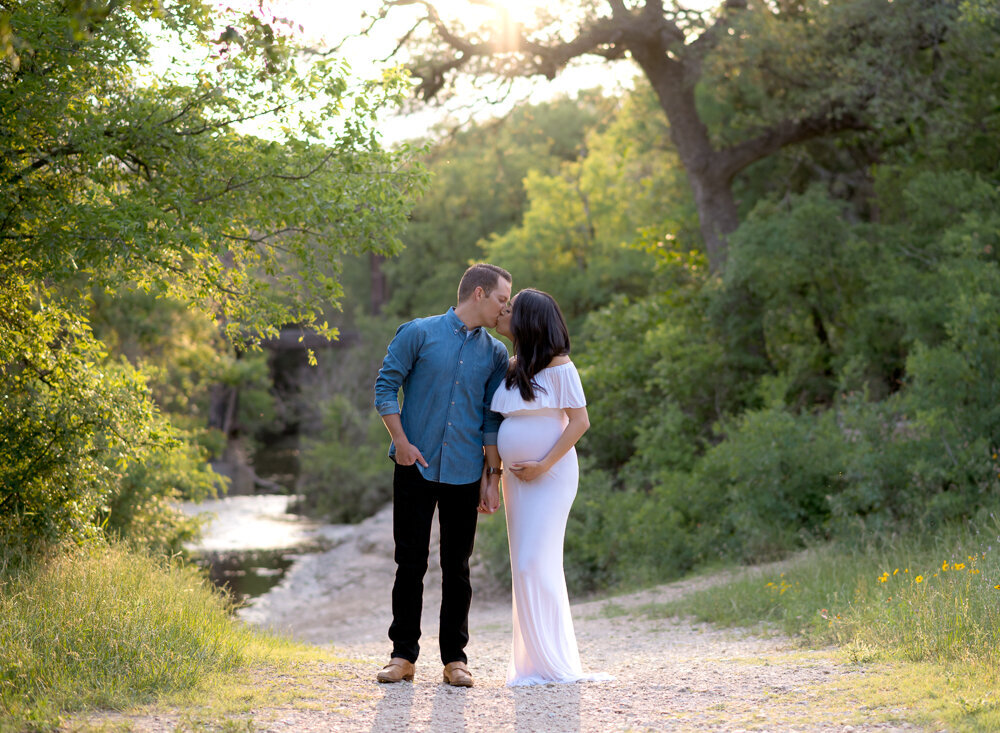 Expecting couple shares a kiss on a wooded trail during golden hour maternity session in Leander, TX