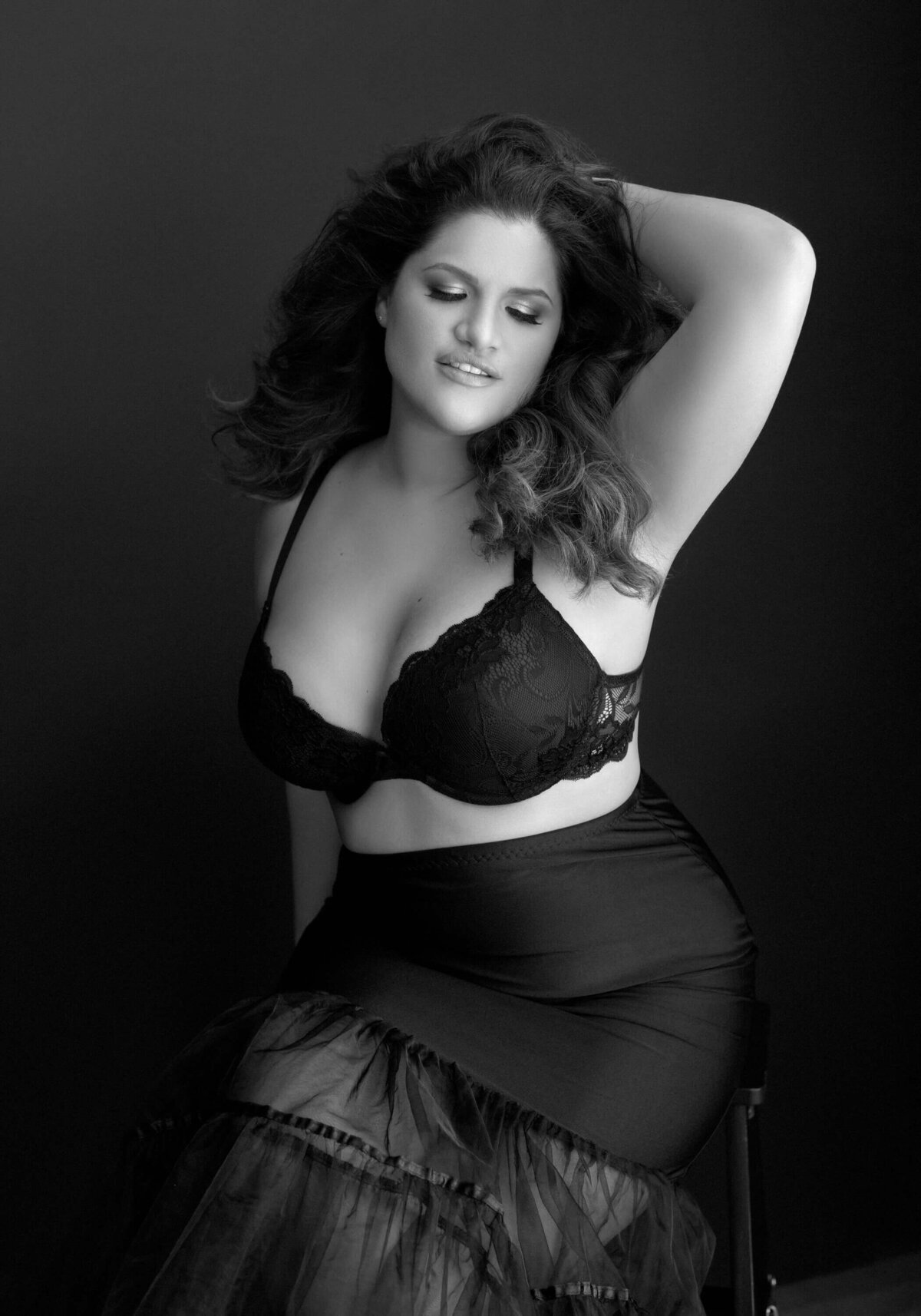 A curvy woman in black lace lingerie and a sheer skirt poses with confidence against a dark background, embracing her beauty and femininity.