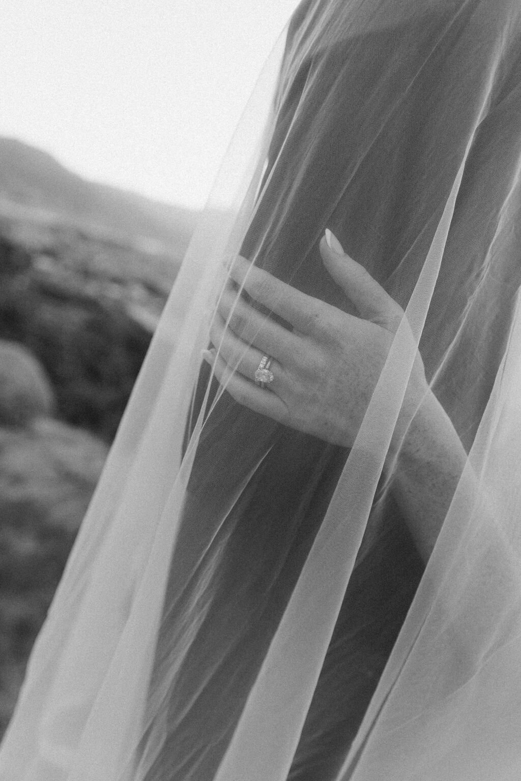 Black and white photo of bride's hand with her wedding ring under veil on groom's arm