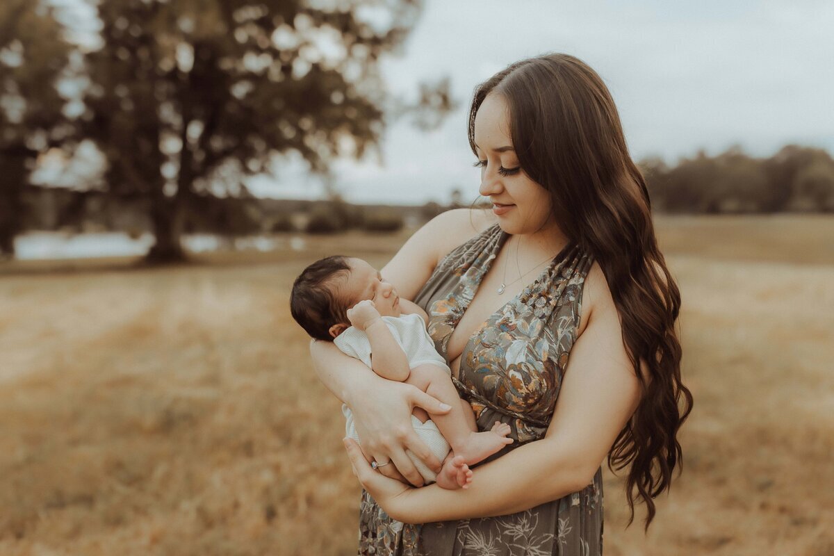 In a serene outdoor setting, a Macon newborn photographer captures the tender moment of a woman with long hair gently holding her baby in her arms amidst the lush grass.