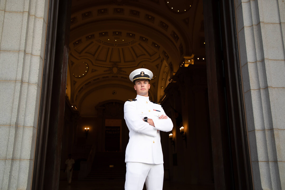 Midshipman senior photography in whites with arms crossed outside Bancroft Hall at the Naval Academy in Annapolis, Maryland.
