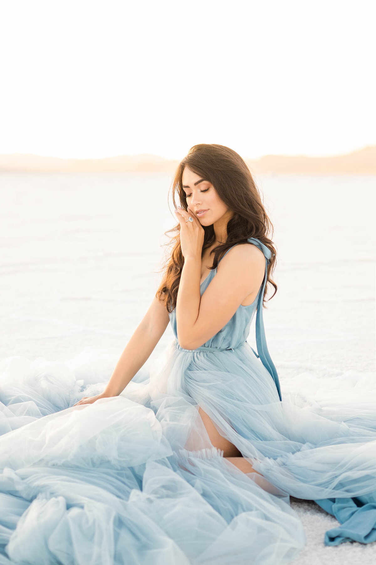 A woman wearing a soft blue gown brushes her hand against her chin while sitting on a white sand dune photographed by Bay area photographer, Light Livin Photography.