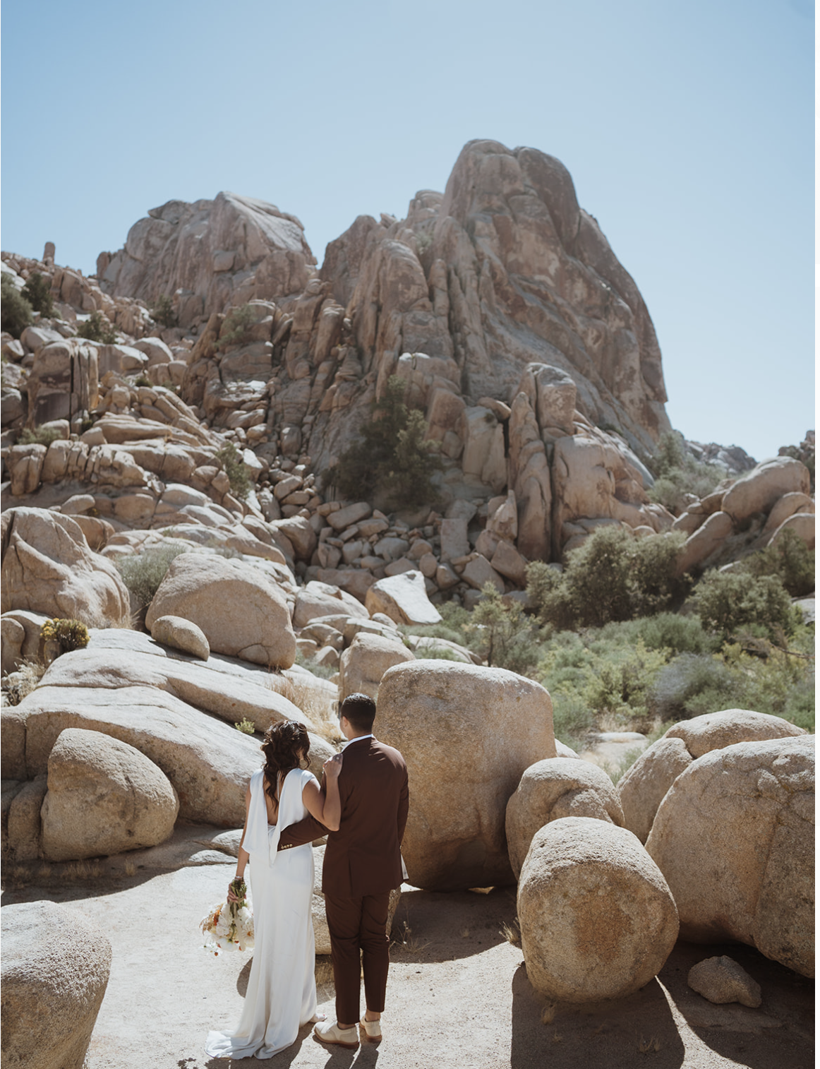 couple is standing looking out into the rocks in joshua tree national park. the groom has his hand around the bride's waist. she has her hand on his shoulder. the groom is in a maroon suit.