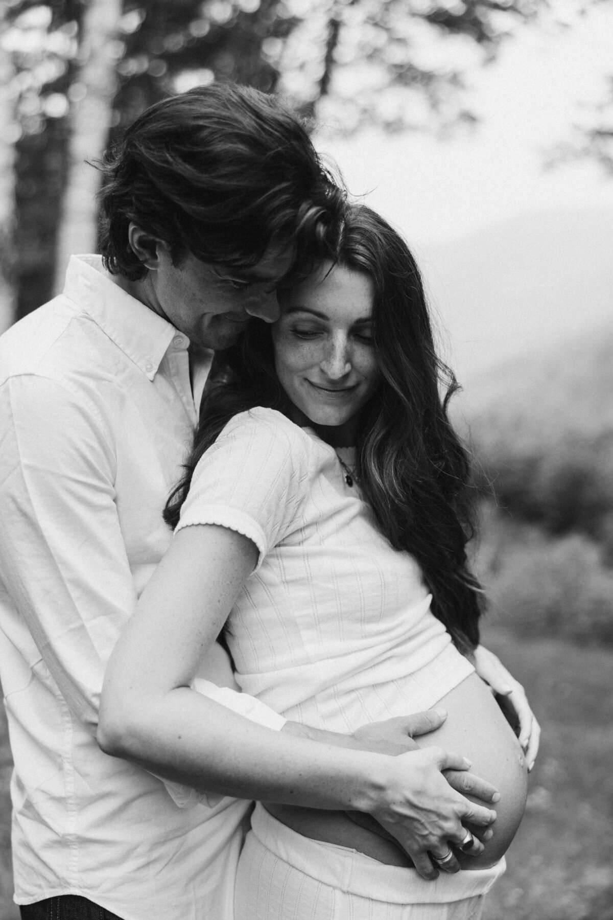 A black and white photo of a pregnant woman and her partner standing outdoors. The man is lovingly embracing her from behind, with his hands gently resting on her belly.