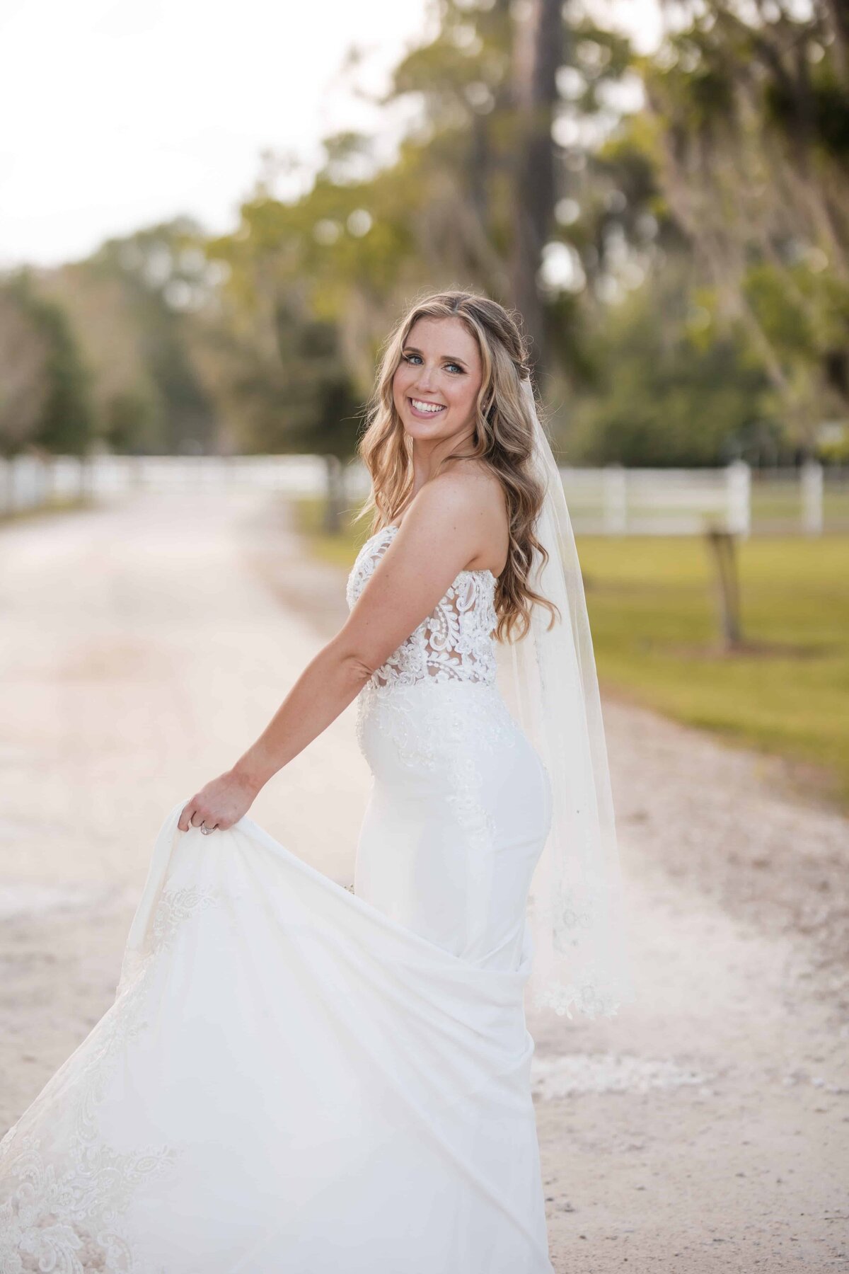 A stunning bride glows with joy as she holds the train of her intricately detailed gown, standing along a sunlit, tree-lined path. Her soft waves, delicate veil, and confident smile exude effortless elegance. The white fencing and lush greenery in the background add a refined countryside charm, blending natural beauty with sophistication. Captured by Phavy Photography in a timeless, true-to-color documentary style that highlights the grace and emotion of the moment—luxury wedding photography rooted in authenticity.