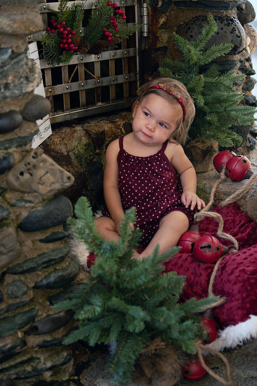 A baby girl sits on a christmas blanket surrounded by tiny trees by a stone fireplace