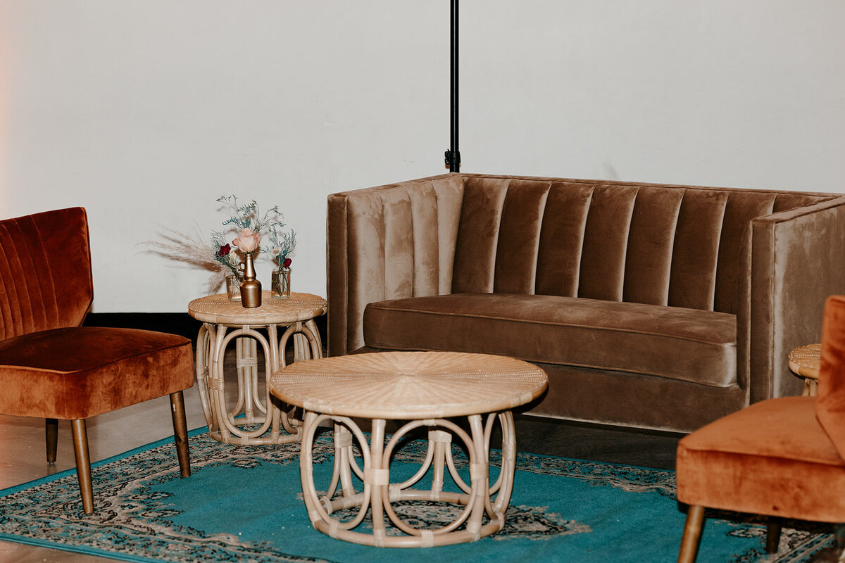 A lounge area featuring a brown velvet couch, rattan coffee and side tables, and orange velvet chairs on a teal rug with floral arrangements in gold vases