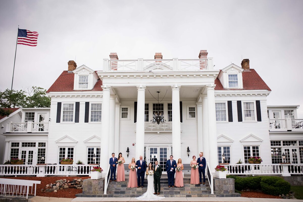 A wide angle shot of a bride and groom standing on the steps of The Manor House surrounded by their wedding party, staggered on the steps on either side.