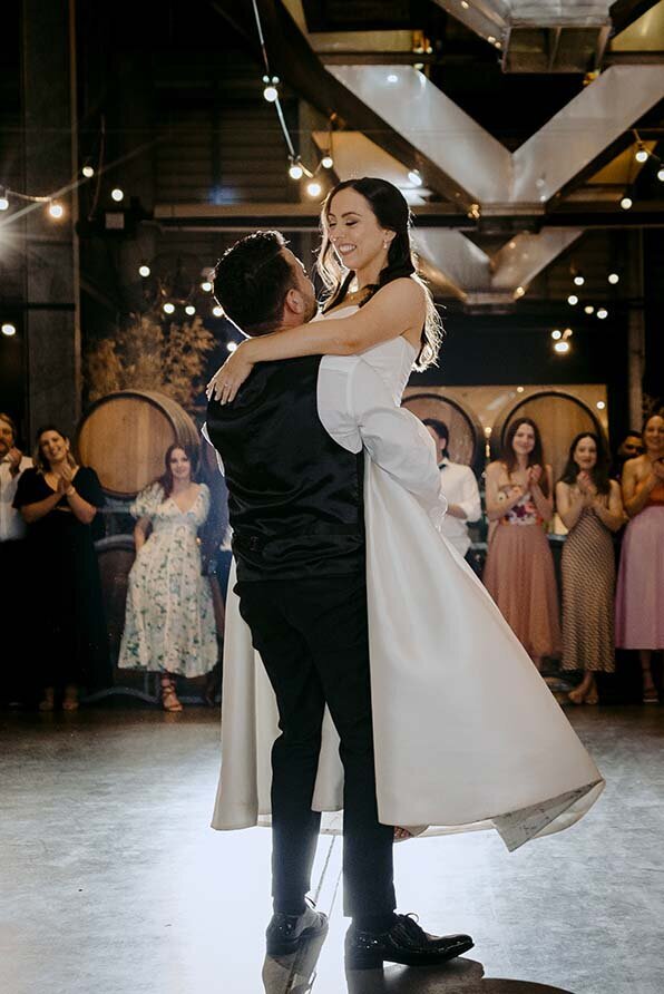 Smiling bride as her partner lifts her in the air during their first dance at a rustic Australian wedding venue with wine barrels and string lights.