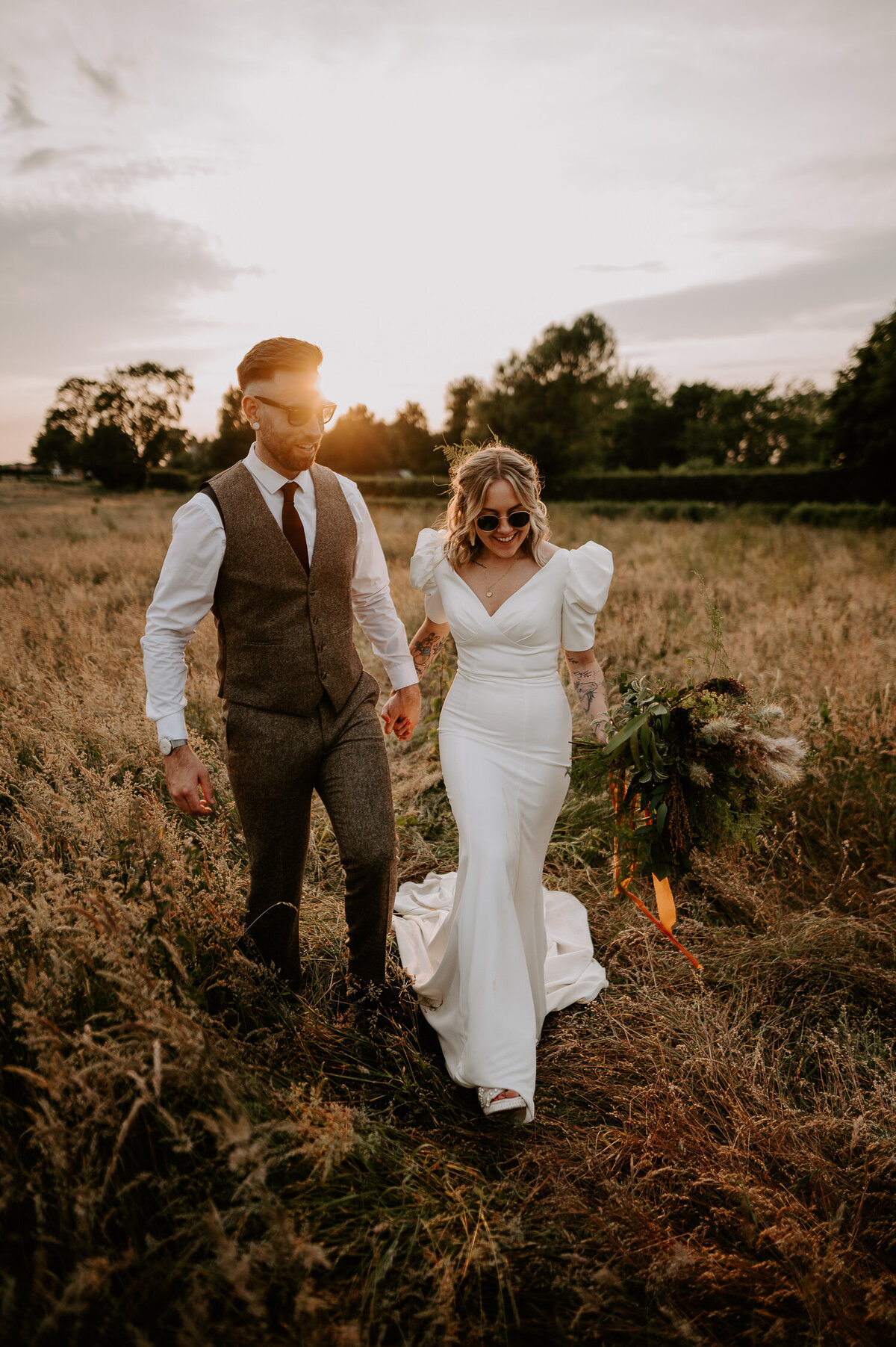 A Bride and Groom walk through the tall grass of the field outside Willow Marsh Farm with their back to an epic sunset, the bride is holding a large wild flower bouquet with pampas grass, her dress is a puffed shoulder fitted dress with a train. The Groom is wearing a brown tweed suit with burgundy tie and burgundy Dr Marten loafers.