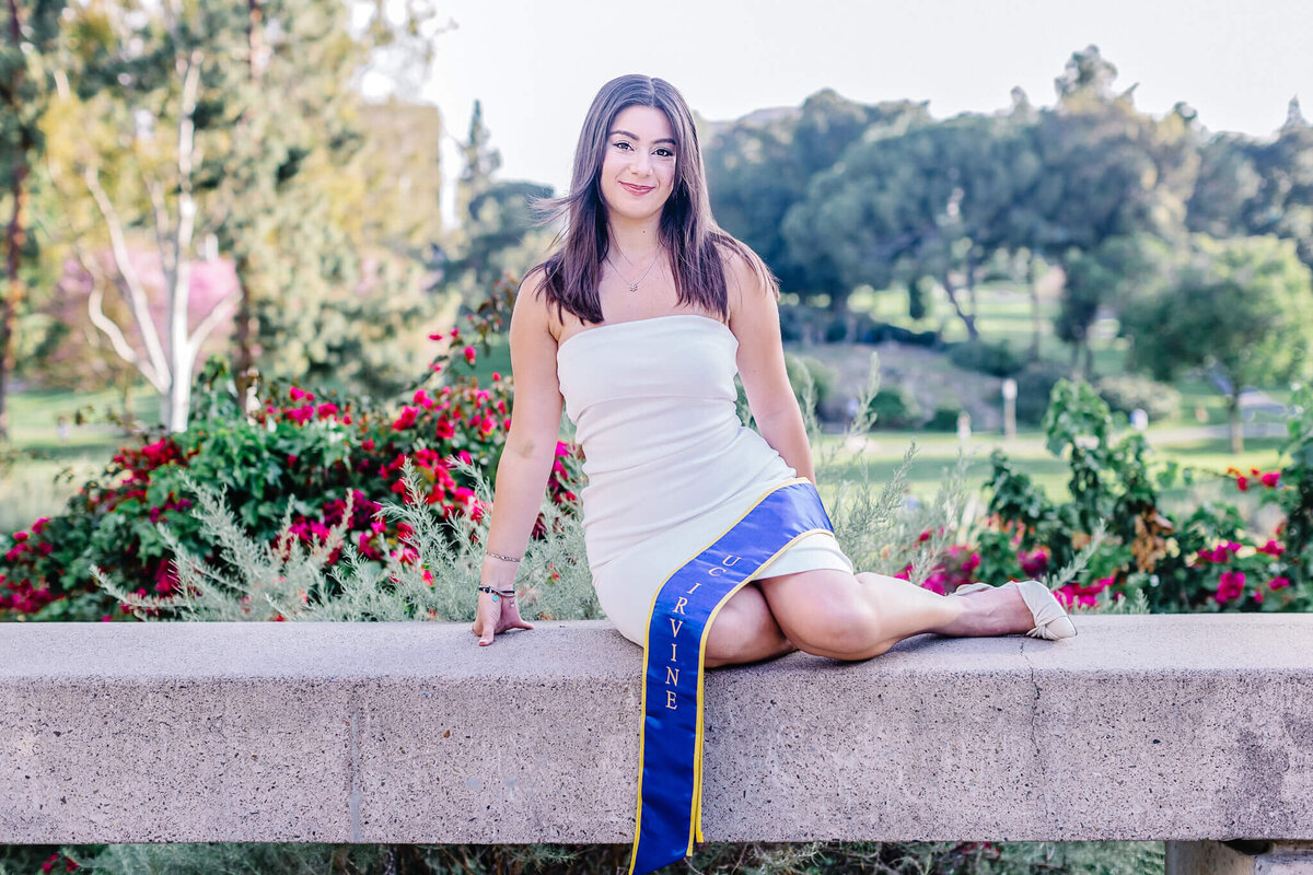 A woman in a white dress sits on a stone ledge in a garden, smiling. Shes wearing a blue and yellow sash with Nineteen written on it. Floral bushes and trees are in the background.