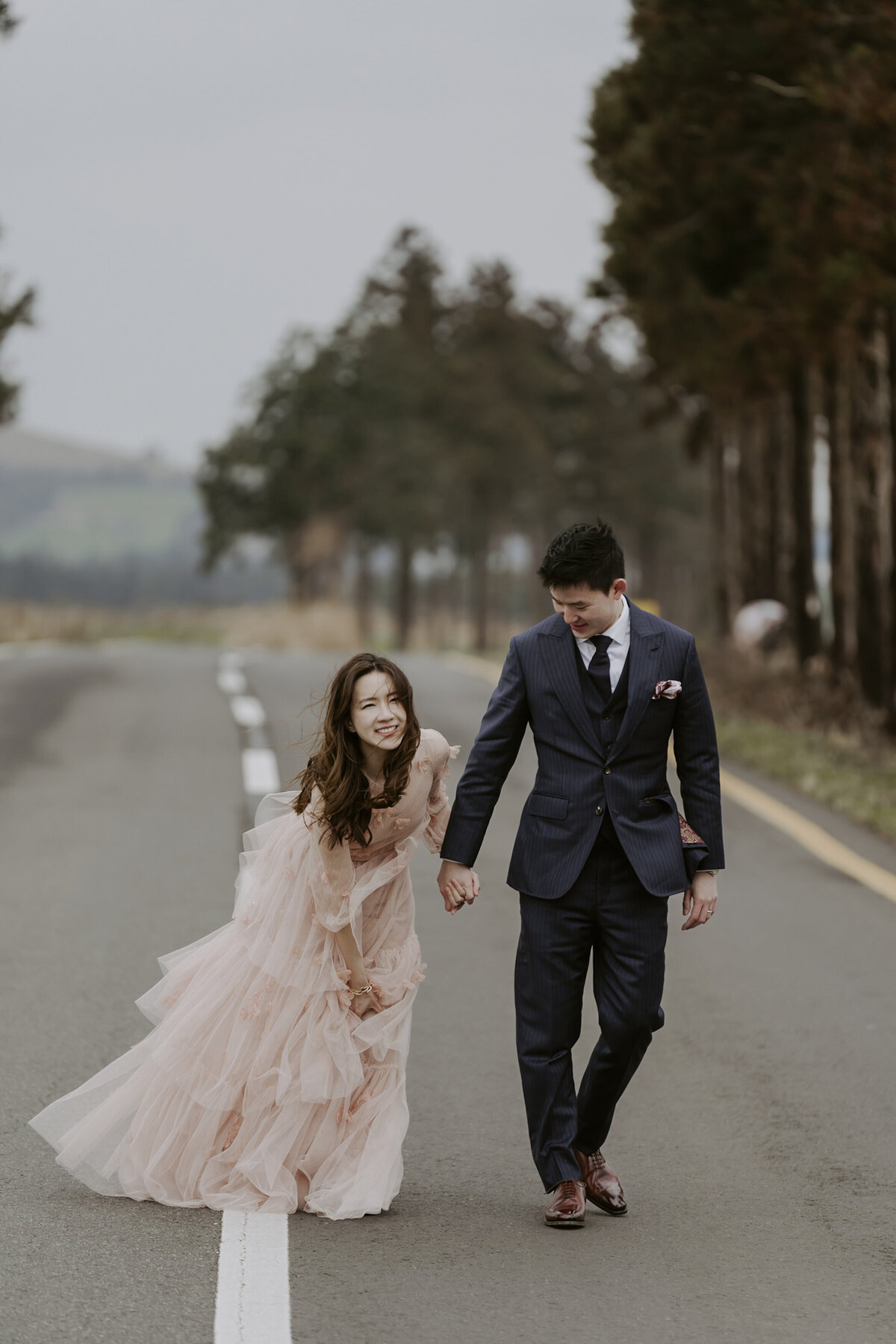 the bride wearing a raffled pink dress while the groom is wearing a black suit and tie while walking in the road of jeju island