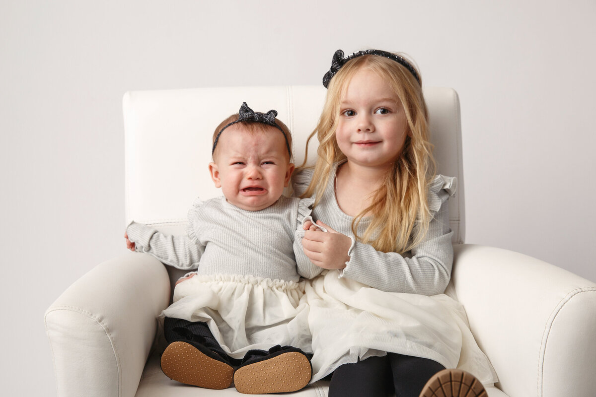 Sisters sitting on a white chair and photographed on a white background.