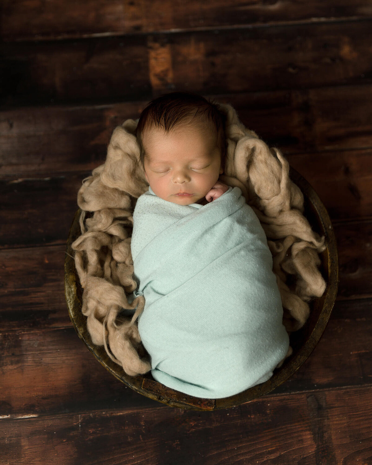 Newborn boy laying in a wooden bowl swaddled in a light blue wrap.