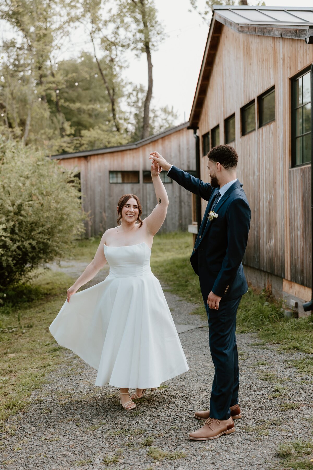 Groom twirls his smiling bride in a tea-length wedding dress during their outdoor portraits at a rustic venue with wooden buildings.