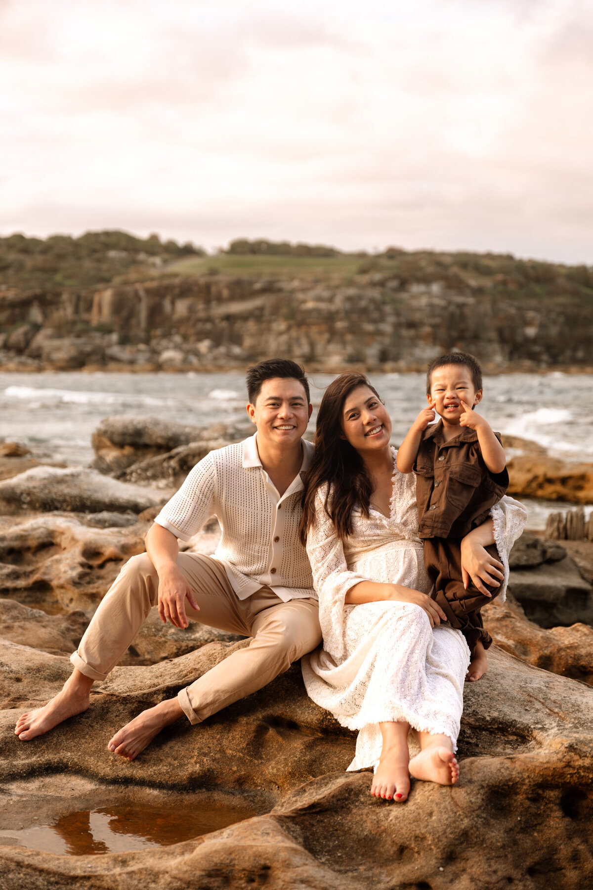 Young family sitting on coastal rocks during a maternity session in Sydney, featuring a smiling couple with their toddler at sunset.