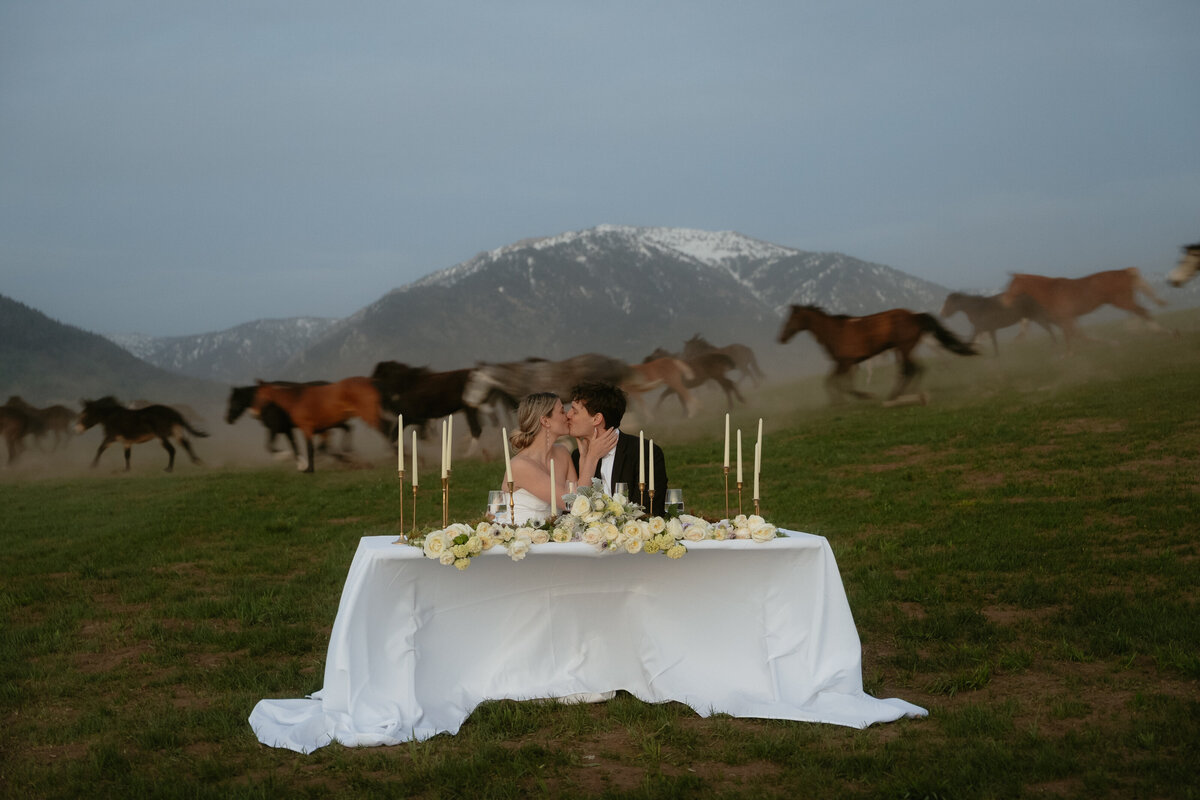 horse herd running in the back as a couple are kissing at their food table with the wyoming mountains in the back