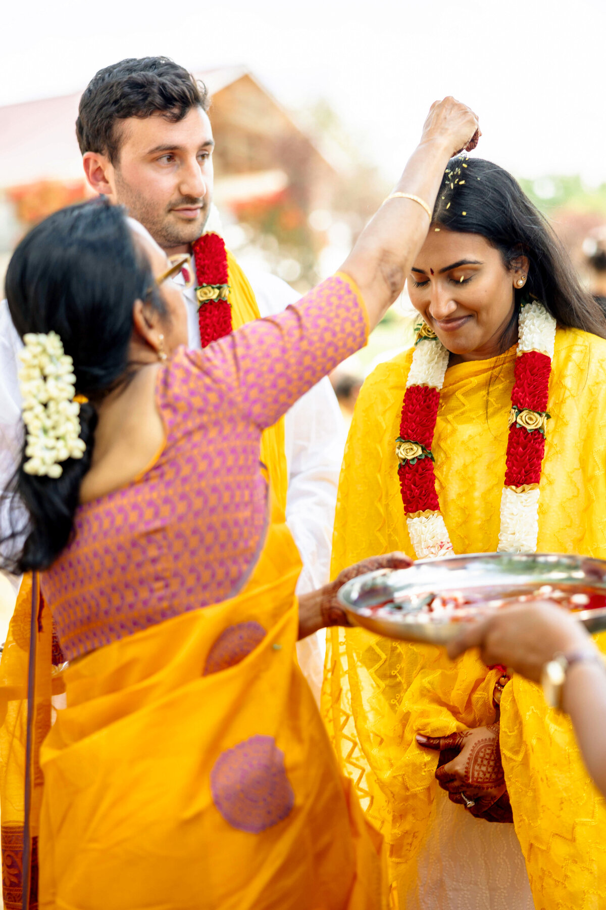 A woman in traditional attire places flowers on a smiling brides head during a ceremonial ritual. The bride, draped in a yellow sari and floral garlands, stands beside a man in a white outfit. A vibrant, cultural setting surrounds them.