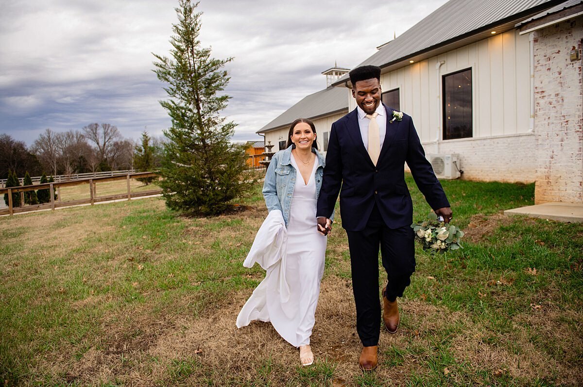 The bride, wearing a long satin wedding gown with a v-neck and and a light blue denim jacket walks hand in hand with the groom wearing a black suit with a white shirt and ivory tie. The groom is holding the bridal bouquet of white, roses, seeded eucalyptus, ivory roses, silver dollar eucalyptus, and white anemone as they walk by the white barn through a field at their winter wedding at Steel Magnolia Barn.