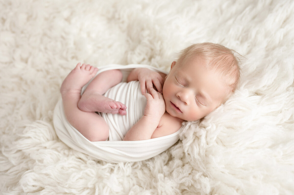 Newborn baby boy swaddled in a white wrap sleeps all curled up on a fuzzy cream blanket during his newborn session with Katie Louise Photography in Bridgeville, PA.