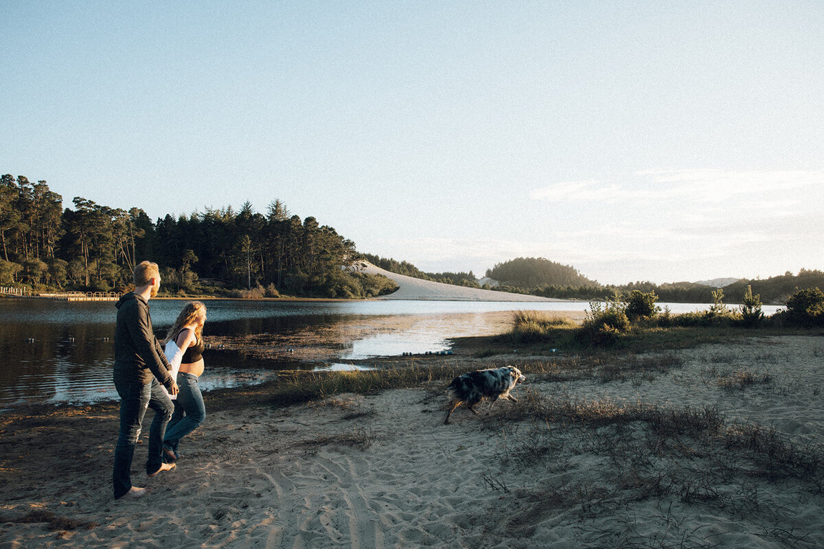 Man and wife hold hands while their dog runs ahead of them on the beach during golden hour on the Oregon coast. 