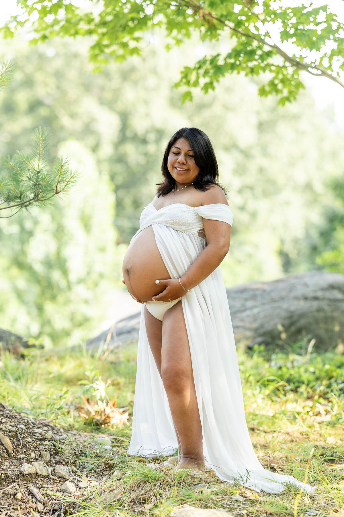Pregnant woman in a open belly white dress is standing on a lush green cliffside backyard during a serene maternity session in Connecticut