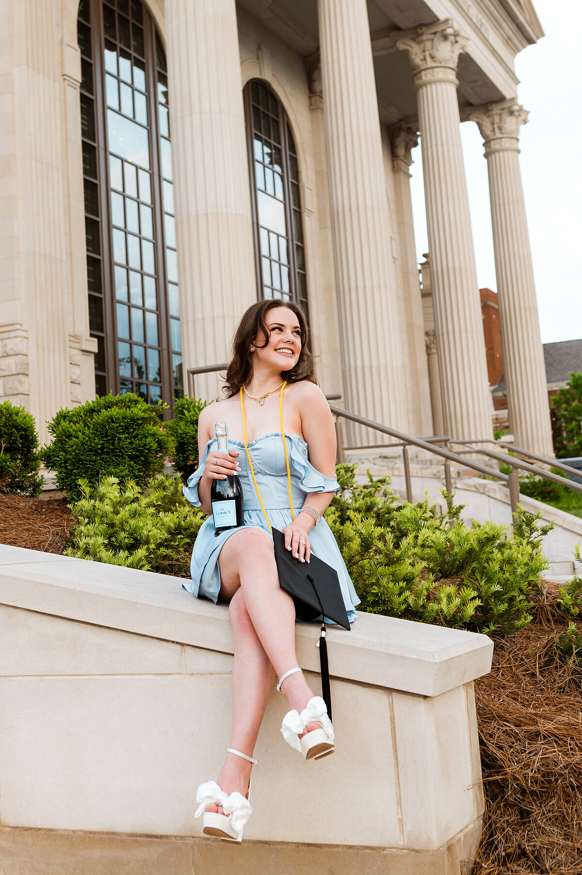 Senior sitting on ledge outside of Belmont fine arts building holding champagne and graduation cap