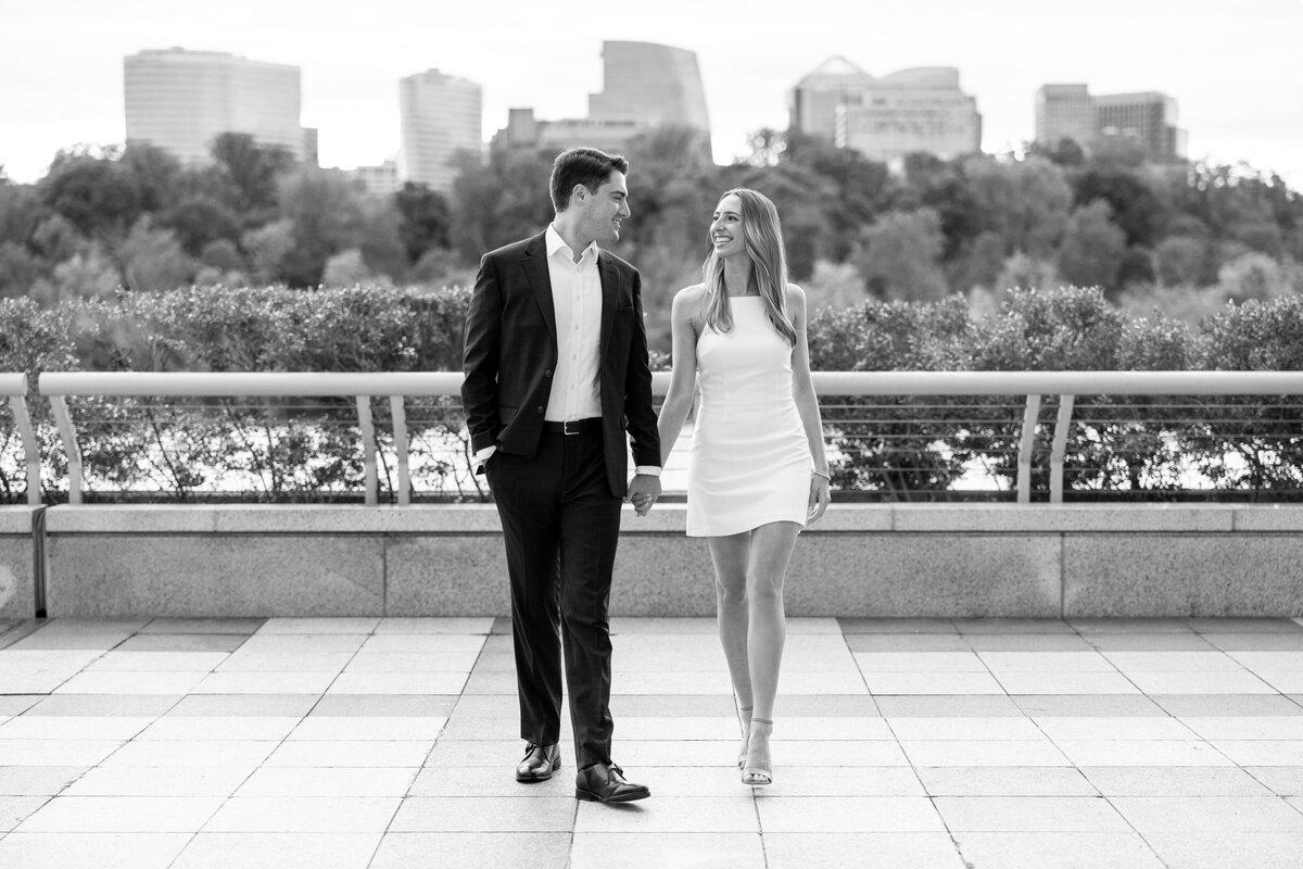A couple walks hand in hand, smiling at each other. The man wears a suit and the woman a white dress. They are strolling on a tiled path with bushes and a city skyline in the background. The image is in black and white.