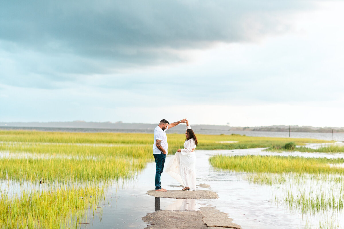 A man is twirling a woman, both smiling at each other. They are standing on a small piece of land, surrounding by shallow water that has long grass poking out from it for miles.